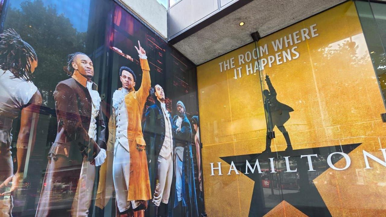 The front of Theatre Royal Plymouth is pictured. It is posters on a large stone building. On the left there is an image of the cast who play Hamilton in action. On the right is a poster promoting the show - with a shadow of a person pointing in the air while stood on top of a star.