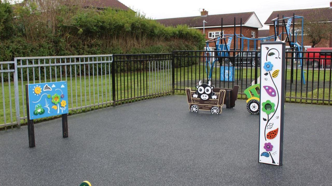 An enclosed play area with smooth tarmac flooring. A rectangular sign with a light blue background, a sun, frog and flowers can be seen on the left. On the right is a long post with colourful flowers on it. In the middle is a cartoon car with a cow in it. You can see grass in the background and a blue climbing frame.