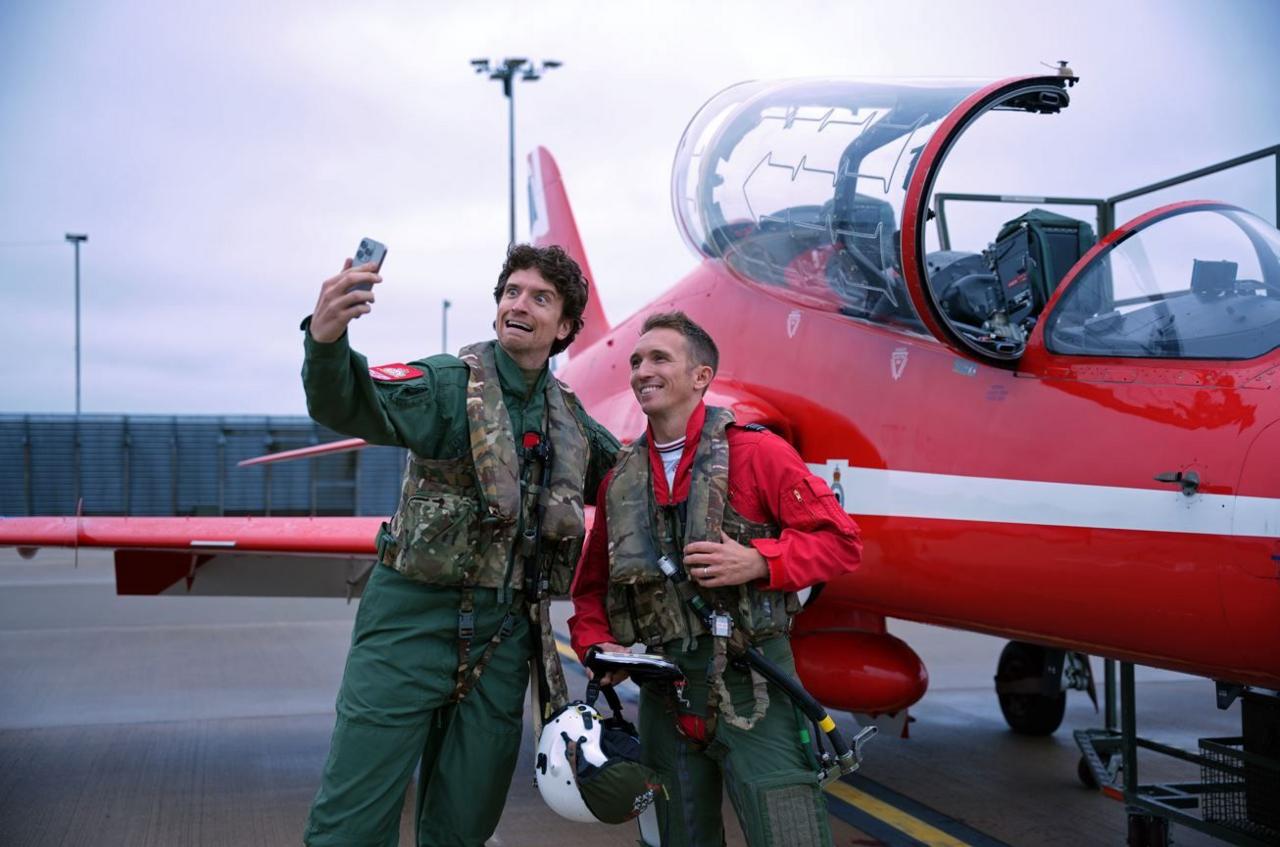 A man pulling a scared face is taking a selfie with another man. They are both wearing overalls and protective camouflaged gear. They are stood next to a Red Arrows plane.