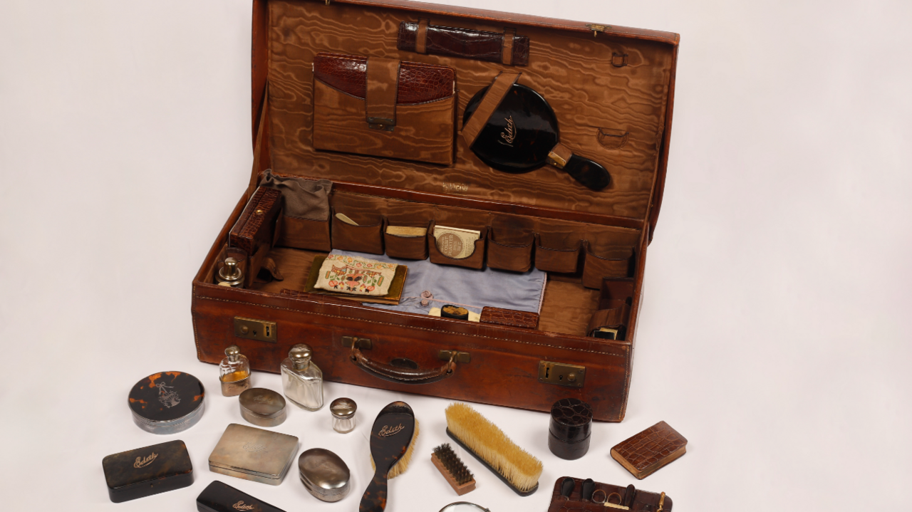 A photo of Mrs Pretty's suitcase. Several items rest inside the case with other items resting outside of it against a white backdrop.