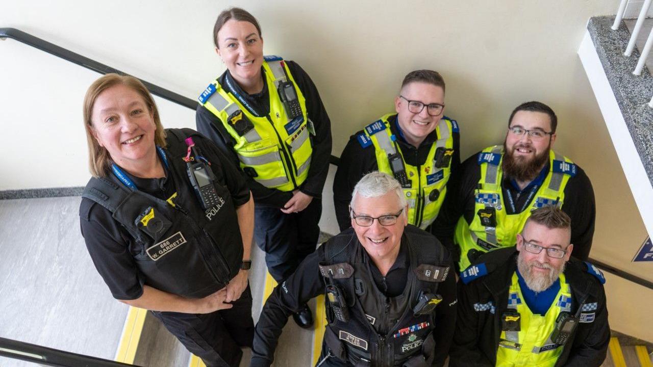 Six police officers are stood, in uniform, on a staircase while looking up towards the camera and smiling.