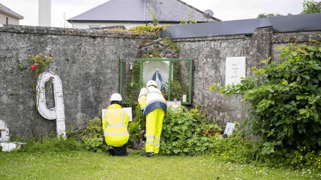 Two workers in yellow high visibility clothing at a memorial left at the site of the former Mother and Baby Institution in Tuam, County Galway.