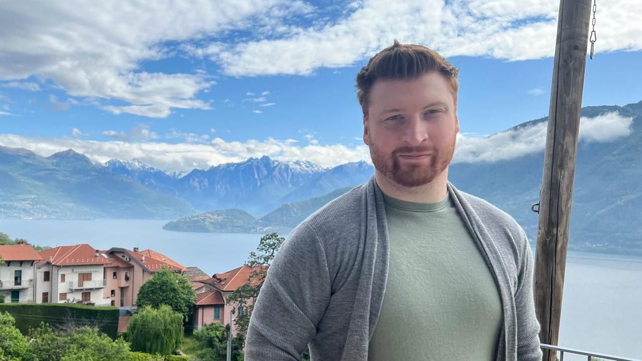 Sam is standing looking at the camera, in the background some buildings and then a large body of water and then mountains under a blue sky with a few clouds