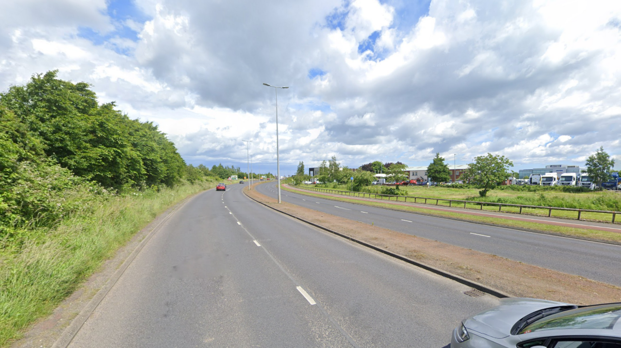 The 1825 way is a dual carriageway with a thin pavement separating the north and south bound sides. Trees are seen on the left of the road. A field, with trucks and industrial buildings behind that, are seen on the right of the field.