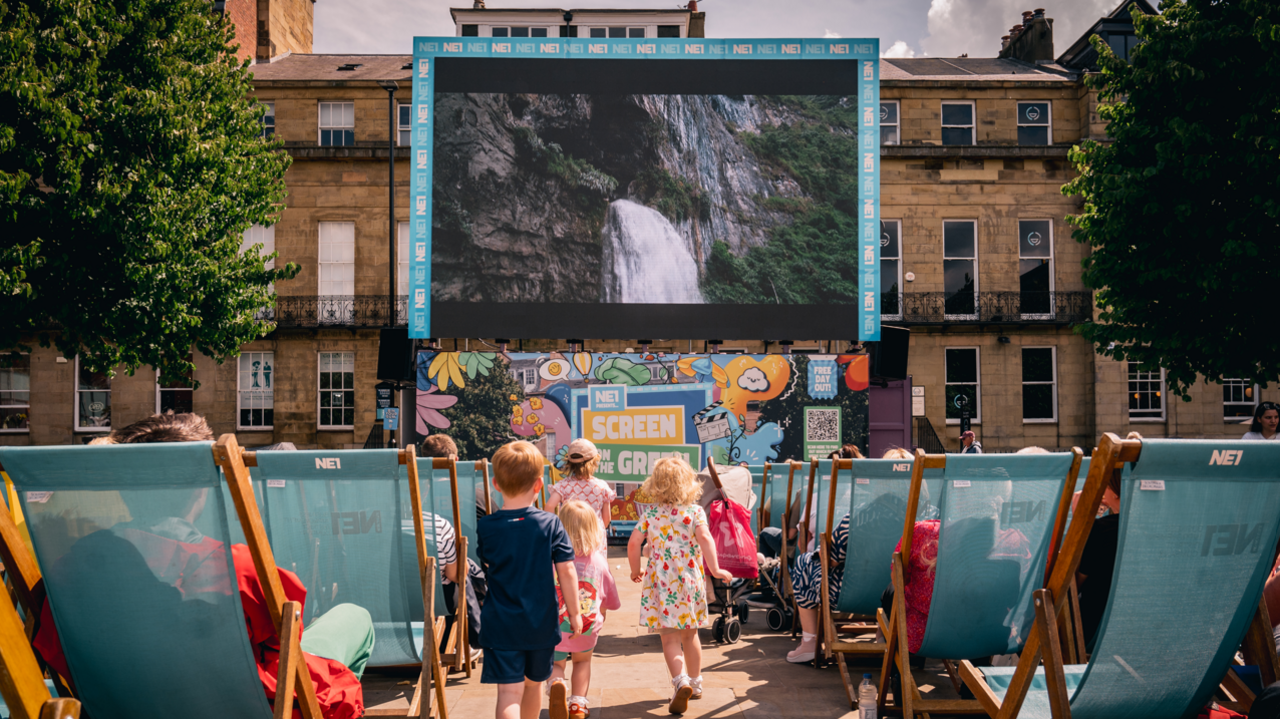 A large screen displaying a picture of a waterfall stands in front a row of buildings in Newcastle's Eldon Square. Pointed at the screen are dozens of blue deckchairs where people are sitting to watch the film. Five children are running between the chairs towards the screen.