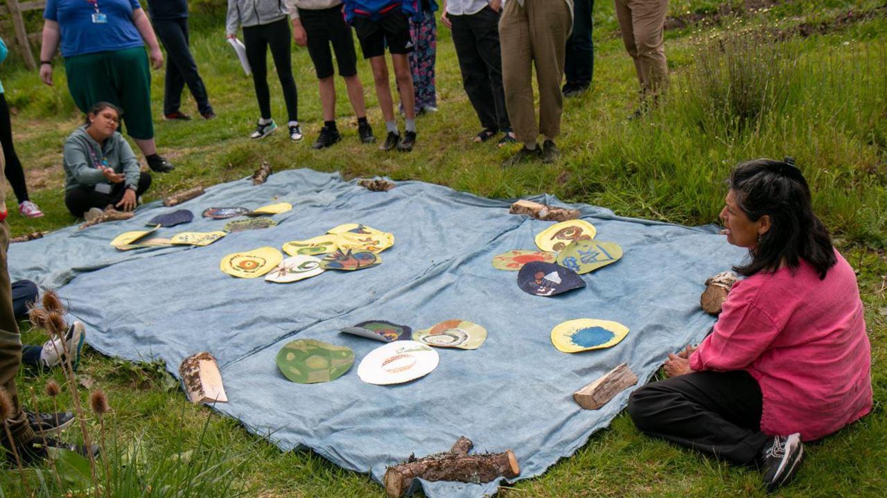 A workshop for the creation of The Consequences by Nature Calling. Participants are outside on a green space. They are working on a canvas with drawings scattered on top of it. One of the participants is sitting down cross-legged while others are standing around it.