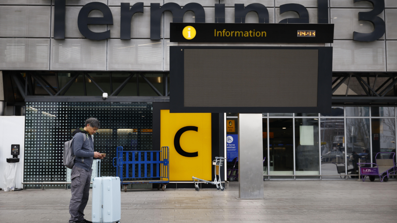 A man stands outside Heathrow Terminal 3, looking at his phone with a suitcase beside him. An empty digital information screen is overhead, and the area is quiet with a few trolleys and barriers visible in the background.