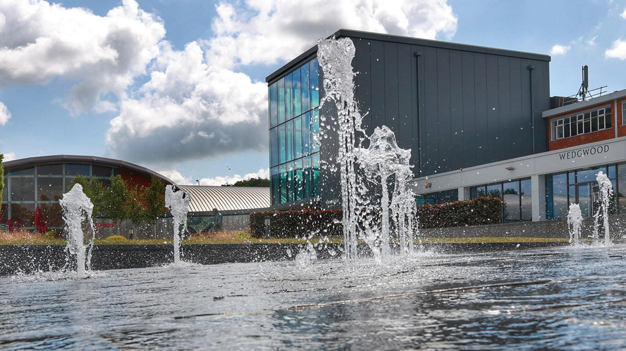 Eight or nine fountains in a shallow pool outside Wedgwood's offices dominate the view. A modern, glass and grey-metal building on the right has a sign outside labelled "Wedgwood". On the left, in the background, is a glass and red rick building with a curved roof