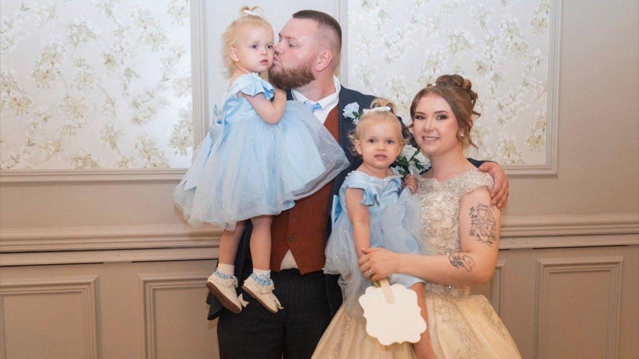 Josh with short hair and a beard kisses the cheek of a young girl he is holding, who is wearing a blue dress. Josh is in a suit and has his arm around Katie, who wears a wedding dress, with her hair up. She is holding another little girl, in a matching blue dress.
