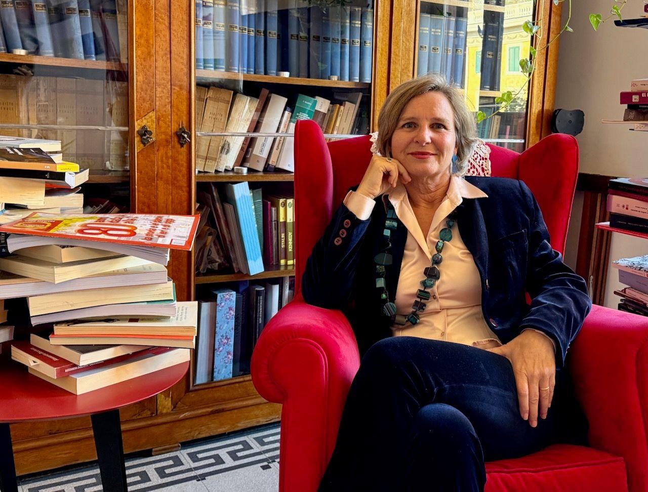 Judge Paola di Nicola sits in an arm chair looking directly into the camera. She is surrounded by books and is wearing a suit and a statement necklace.
