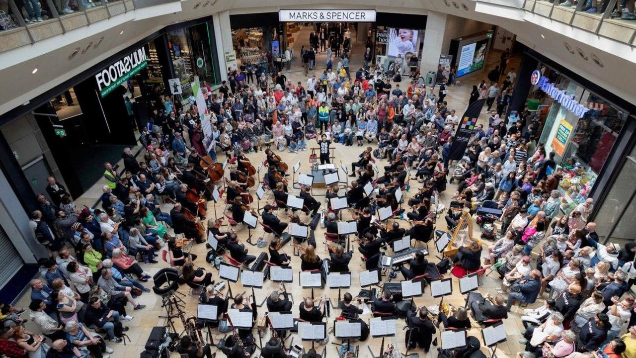Looking down on a shopping centre floor, with a large orchestra gathered in the middle of the space, and an audience on three sides.