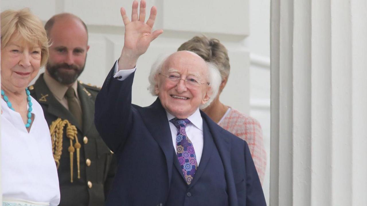 Michael D Higgins is an older man, with white hair placed around his head. He is waving and is smiling. He is wearing a blue three-piece suit, a flower pattern tie and a white collared shirt. Standing beside him is his wife Sabina and an aide behind him. He is stood outside the Áras - which is a white building with large colums.