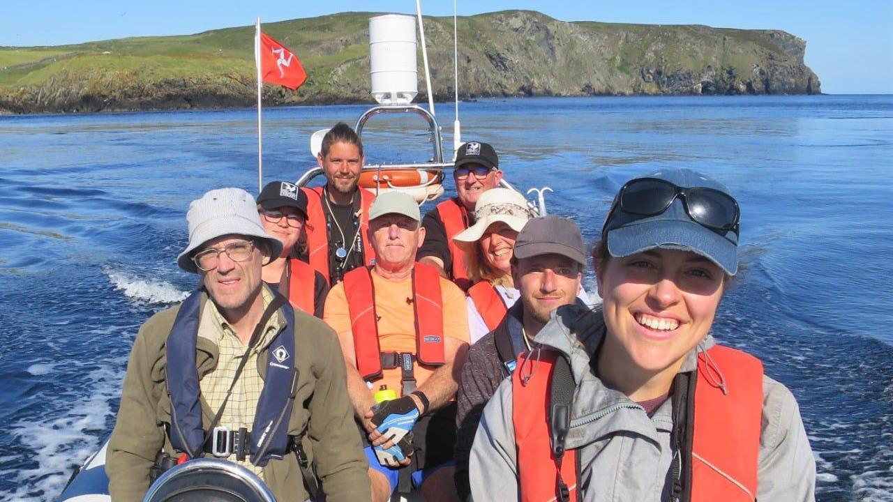 Eight people sitting on a small boat. They are all wear orange lifejackets, and there is a Manx flag flying at the back of the boat, and the Calf of Man in the background.