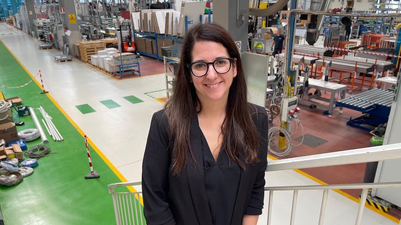 Melania, a woman with long dark hair and glasses, is seen in front of a factory floor