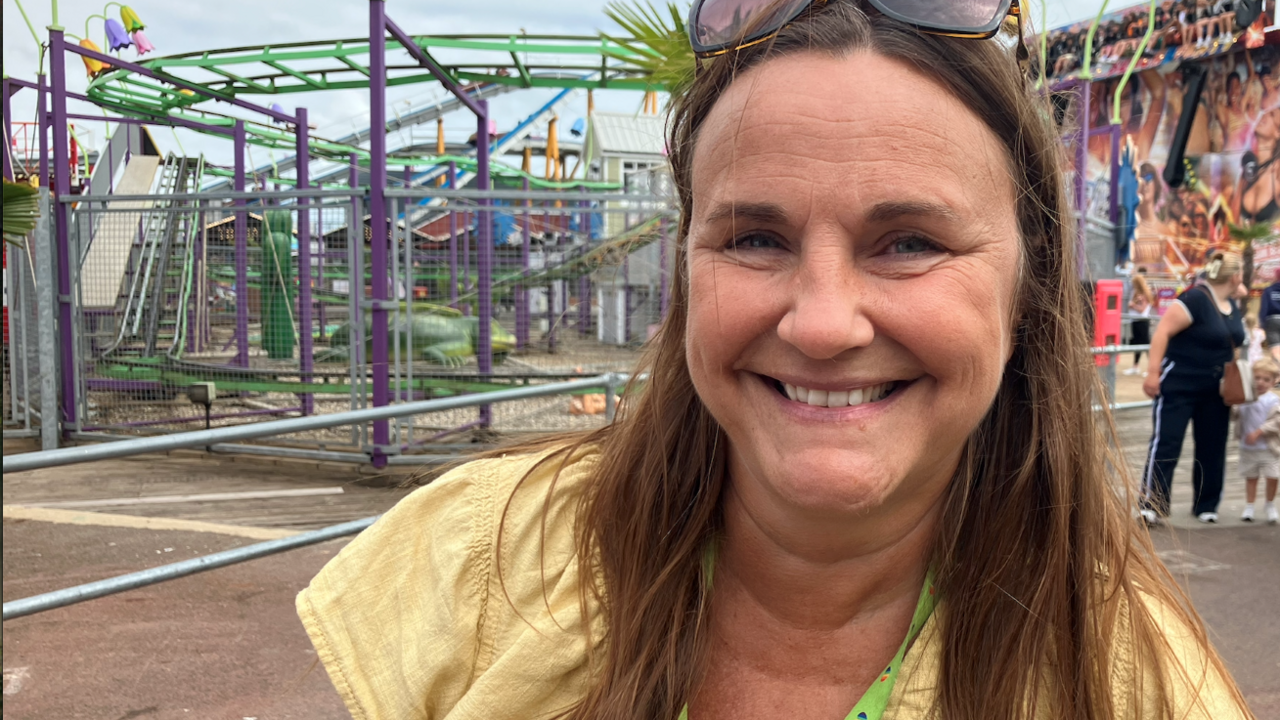 Woman with sunglasses and a yellow top smiles at the camera. Behind her is a fairground ride.