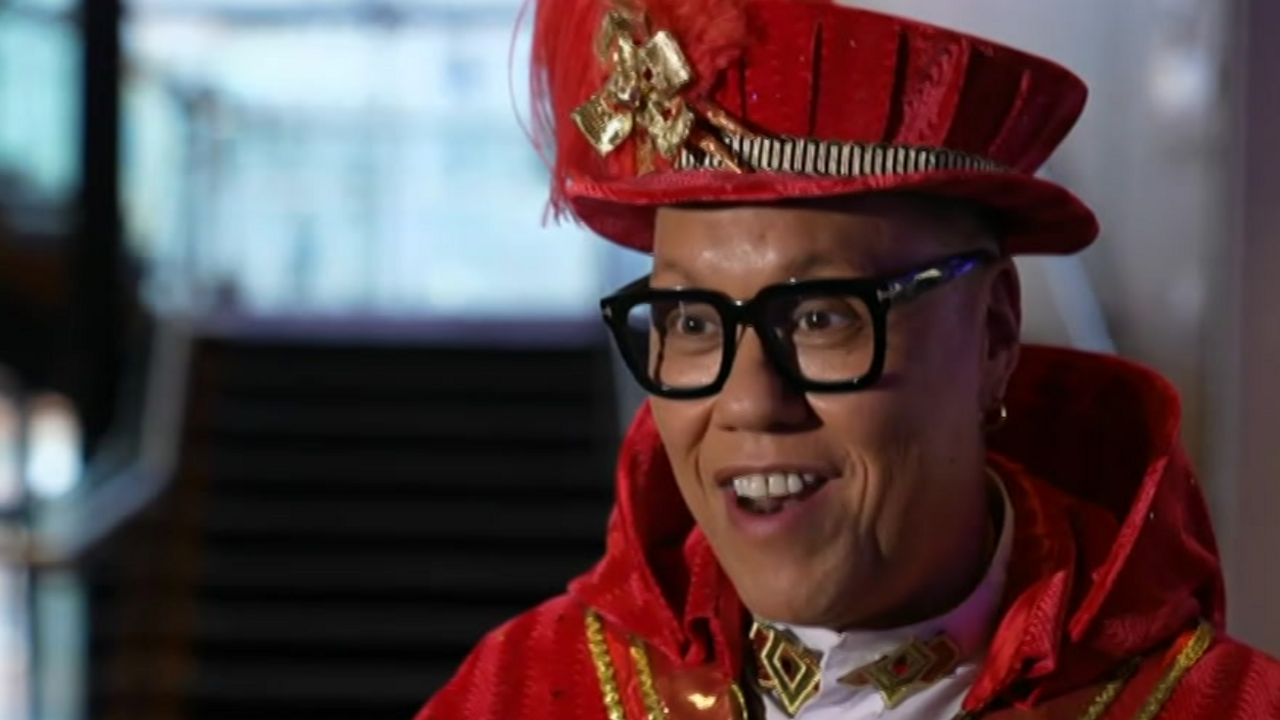 A man in thick-rimmed glasses has an ornate red hat on his head. He is also wearing a red caped top with gold braid on it.