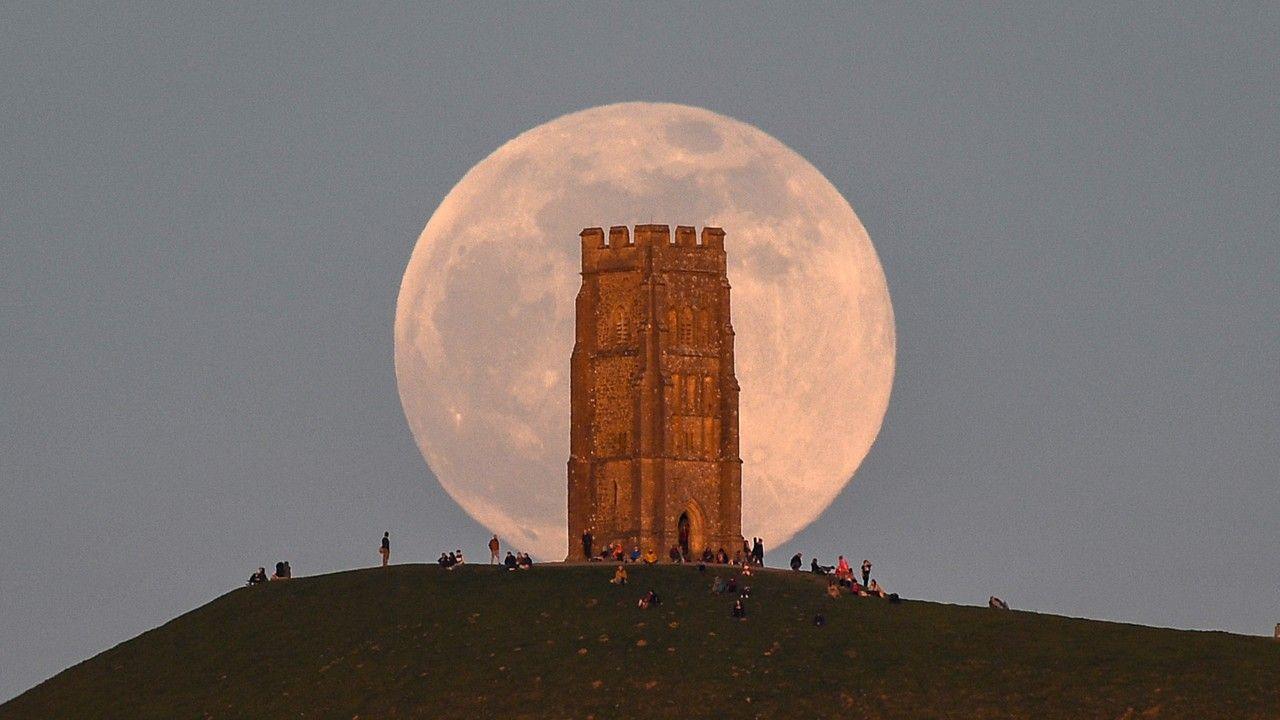 A huge pink hued Moon sits in a grey sky, behind the tower on Glastonbury Tor with people milling around at the base