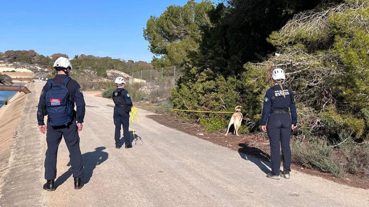 Three individuals have their back to the camera. They all have on Navy overalls and one of them holds a golden Labrador on a lead. To their left is a blue river. To their right is green shrubs. 