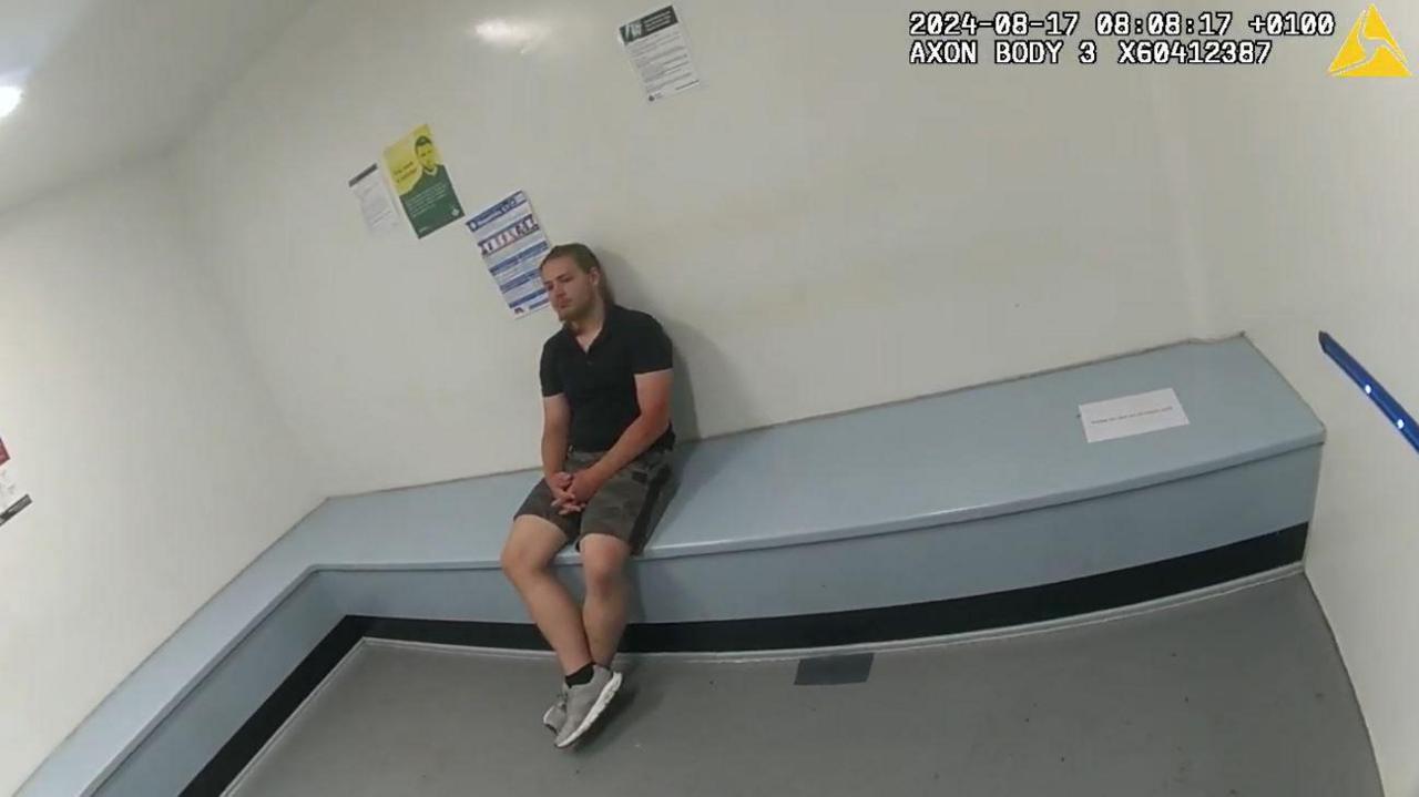 A man sits in a police cell. The room is bare and white. 