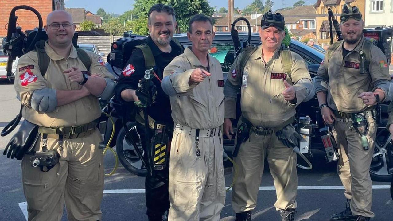 There are five men standing in a row in a car park. They are wearing all-in-one Ghostbusters suits with proton packs on their backs and are smiling at the camera. 