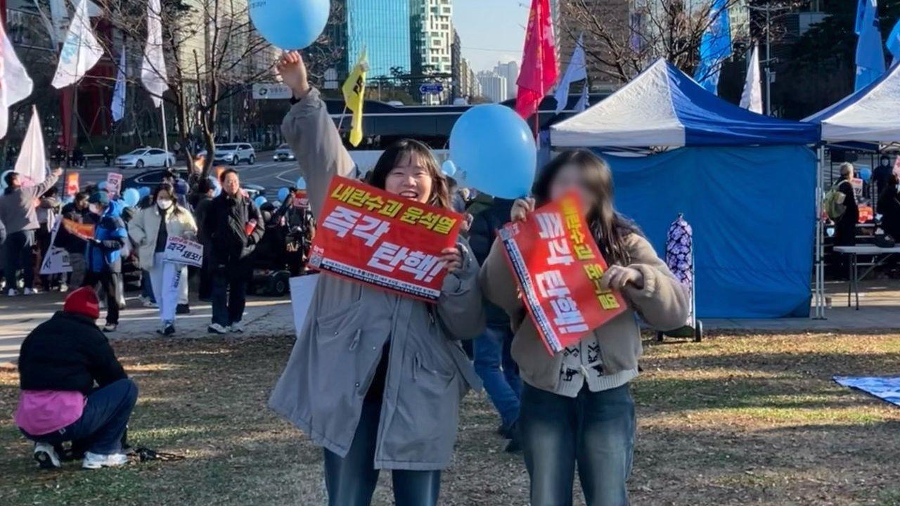 Lee Jinhaa seen holding a red poster with white writing on it calling for Yoon's impeachment. She is waving a blue balloon in the air, while her friend stood beside her is also holding a blue balloon.