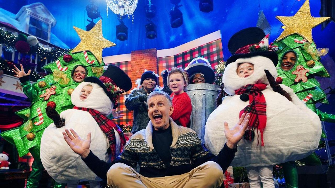 Patrick Kilety pictured with his legs crossed and a number of children in elaborate costumes surrounding him.