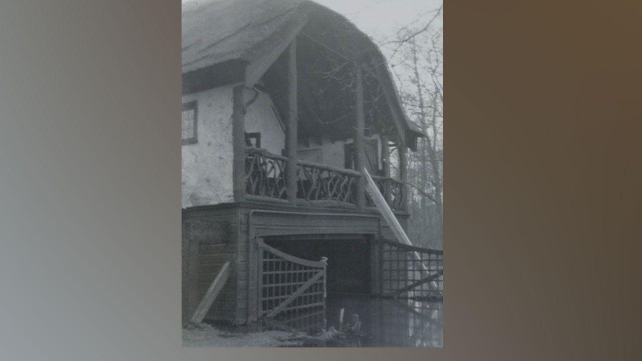 A cropped close-up of a black-and-white showing Staithcote in Wroxham. It is a boathouse built and then extended with a chalet above in the early 20th Century. It shows a two story structure with a thatched roof, a veranda on the first floor with a river flowing underneath the building