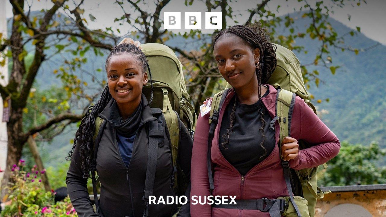 Sisters Elizabeth and Letitia smile for the camera as they take part in Race Across the World. They each carry light green back packs and wooded mountains can be seen in the back ground.