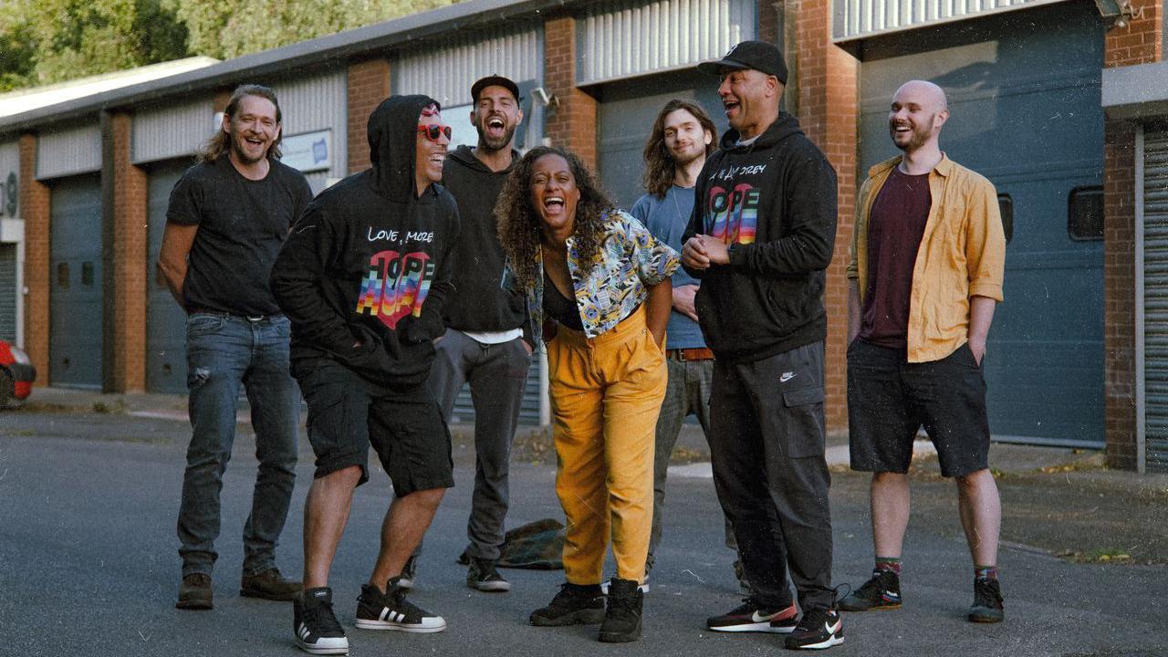 Seven musicians laughing in front of a row of industrial roller doors.