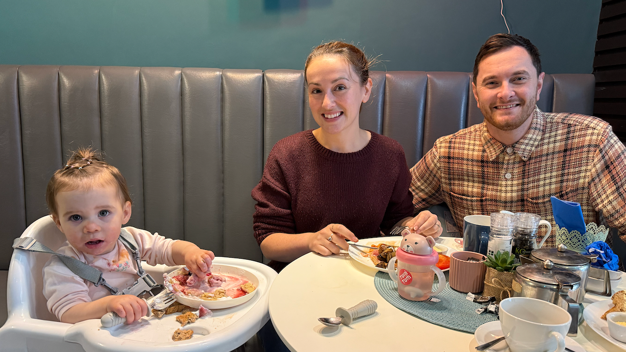 Jane and Adam Petrie, and their daughter Eden. Mr and Mrs Petrie are sitting at a round, white table and Eden is in a white highchair. Eden has a fork in one hand and her other hand in her food. The other table has cups, plates and metal tea pots on it, along with a bear sippy cup.
