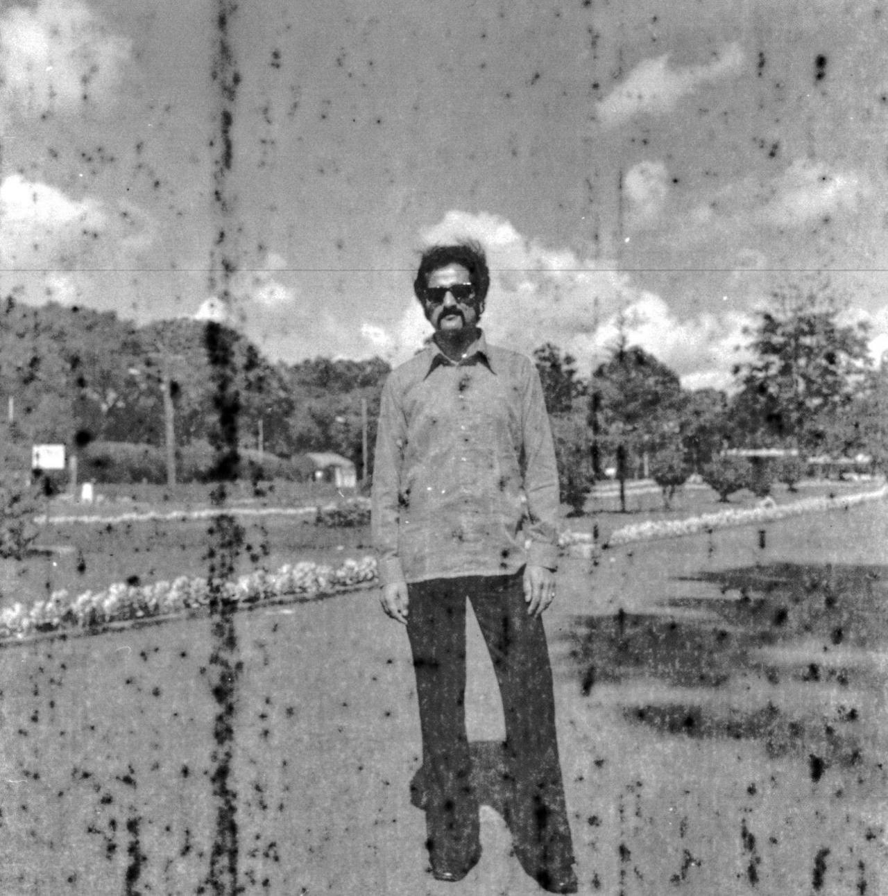 A black-and-white photo of Anil. He has a moustache and is wearing a shirt and sunglasses. He is posing in front of a garden with flowers and trees. The photo shows signs of damage with black spots.