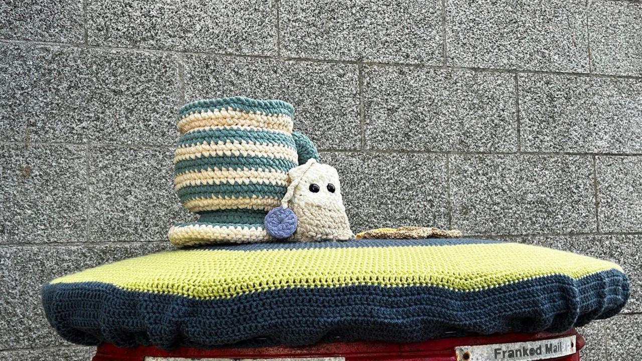 A yellow and black postbox topper with a knitted blue and white mug and knitted white and grey teabag. Behind them is a grey brick wall. 

