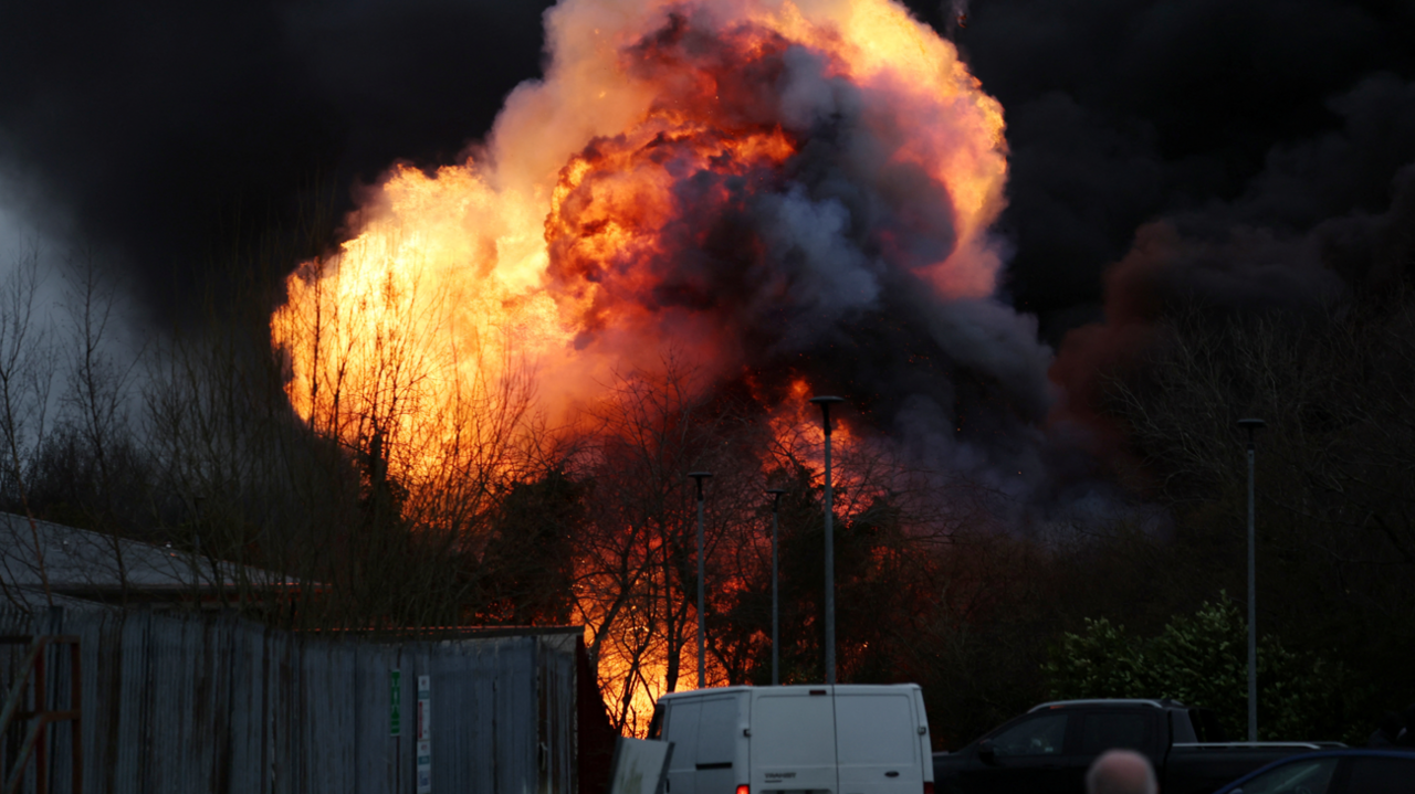 A huge fireball erupts in the sky behind some trees and metal railings. Traffic is parked in the foreground looking on.