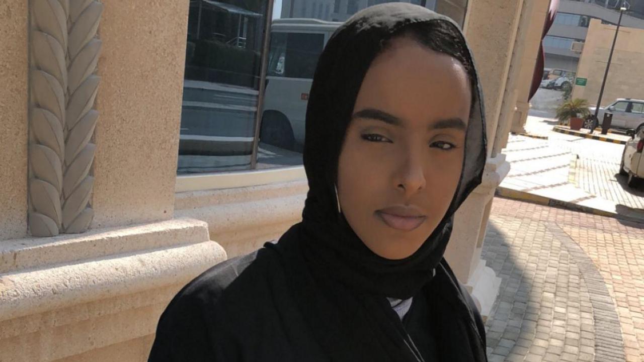 Marjama Osman is seen in a black hijab and abaya standing outdoors, looking directly at the camera in bright sunlight.