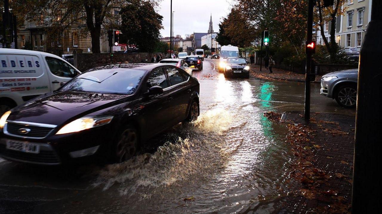 A car driving through a large puddle on a road. It's dark and has been raining. There are other cars in the distance.