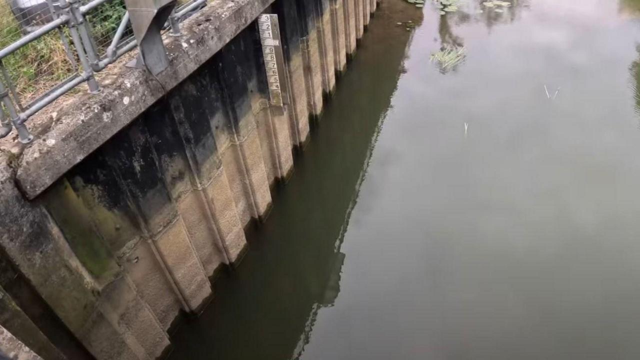 A picture taken from over the river, pointing down at the water below. There is a marker alongside the water which shows the water level has fallen. 