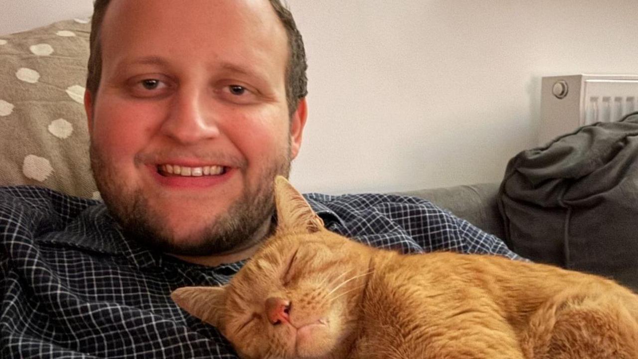 A smiling man in a checked shirt with a sleeping ginger cat on his chest