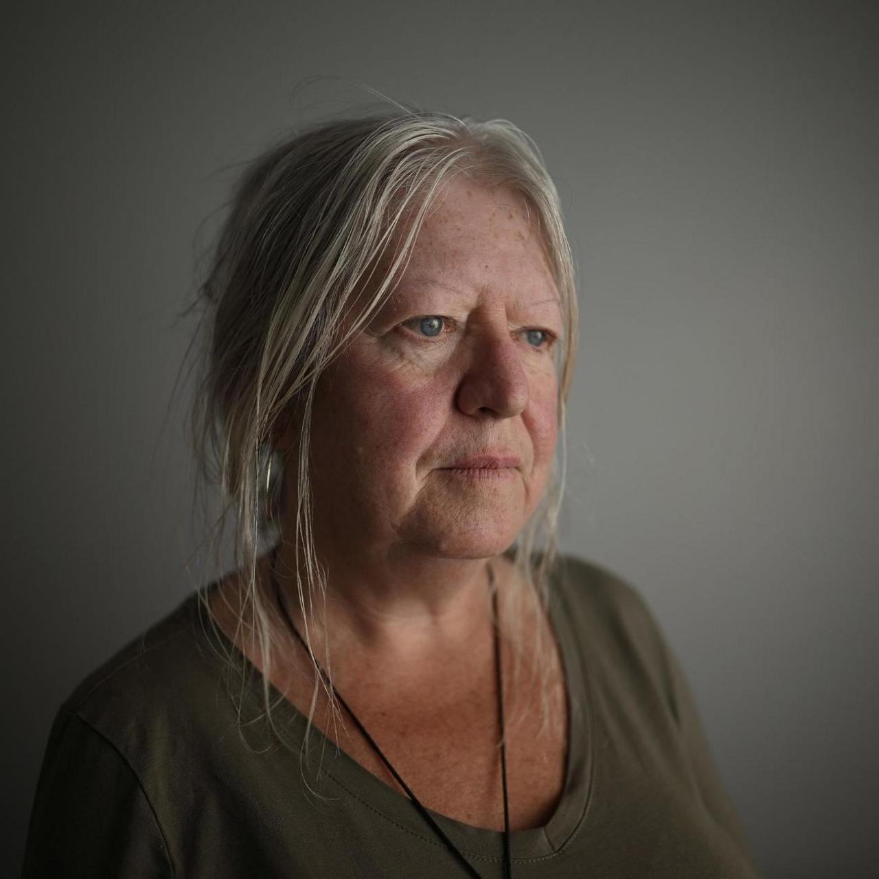 A woman with shoulder-length blonde-grey hair looks off to the side. She is standing in front of a light-coloured wall. She is wearing an olive-coloured top.