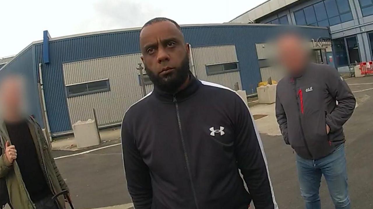 A still from police bodyworn camera footage, showing Israar Shah wearing a black branded sports zip-up jumper, facing towards the camera. Two other people stand near him, not in uniform, with their faces blurred. A large flat-roof warehouse building is in the background.