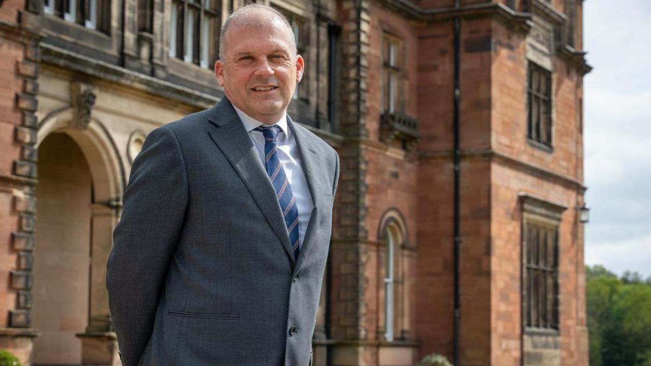 Prof Kevin Shakesheff is wearing a blue striped tie, white shirt and grey suit jacket. He is looking at the camera and standing in front of a building.