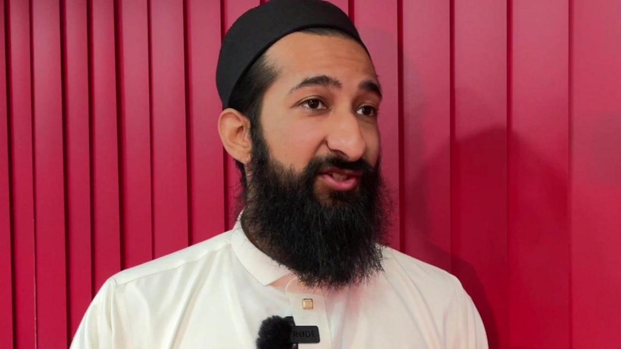 A man with dark hair and a beard is stood in front of a red screen. He is wearing a dark hat and white shirt.