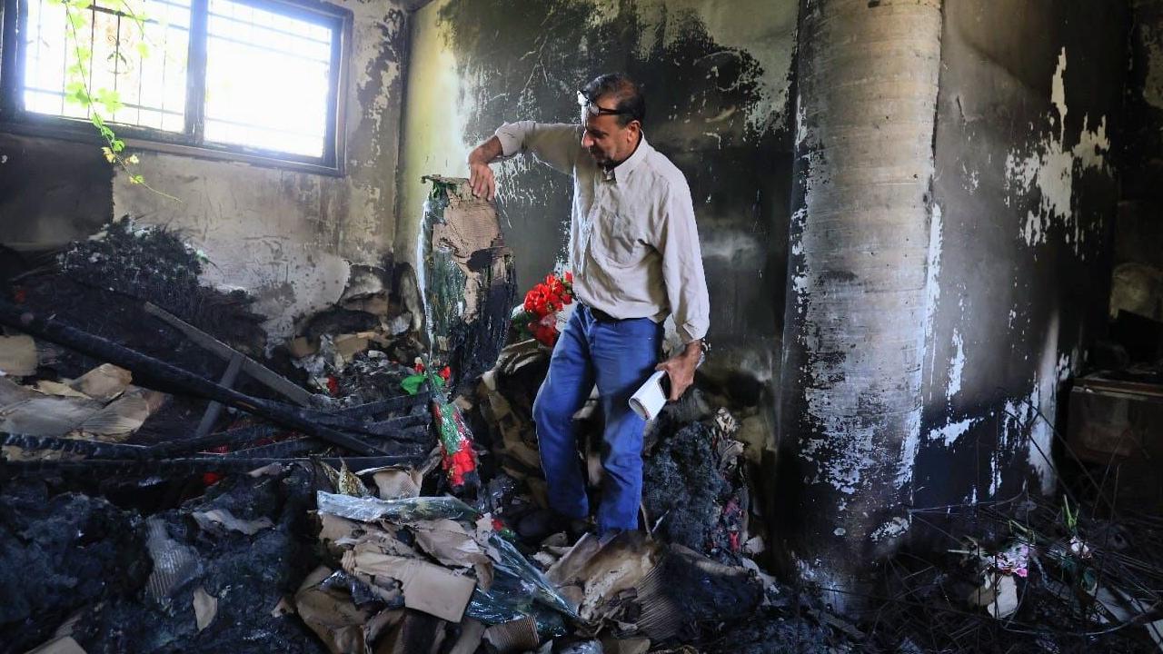A man in a pale shirt and jeans stands in a room blackened by fire holding burnt items including colourful artificial flowers.