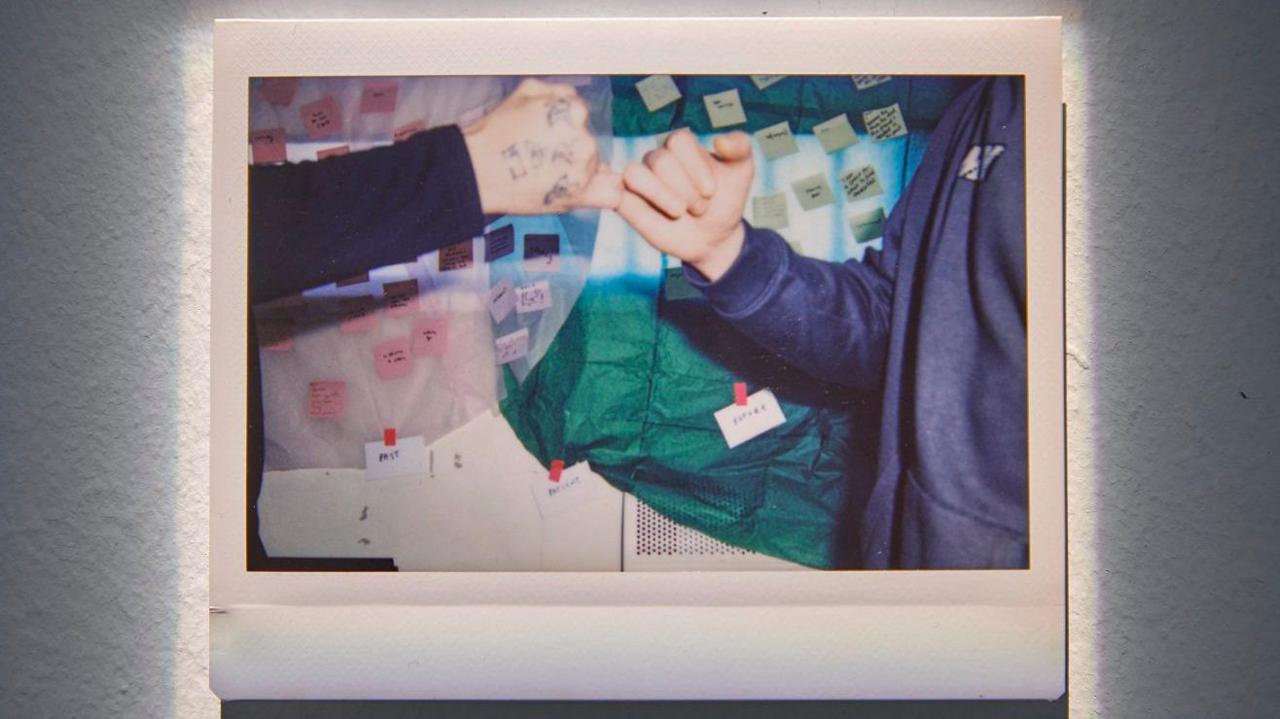 A photo of two people's hands reaching out to 'fist bump'. In the background are fabrics and mini Post-It notes on a wall. 