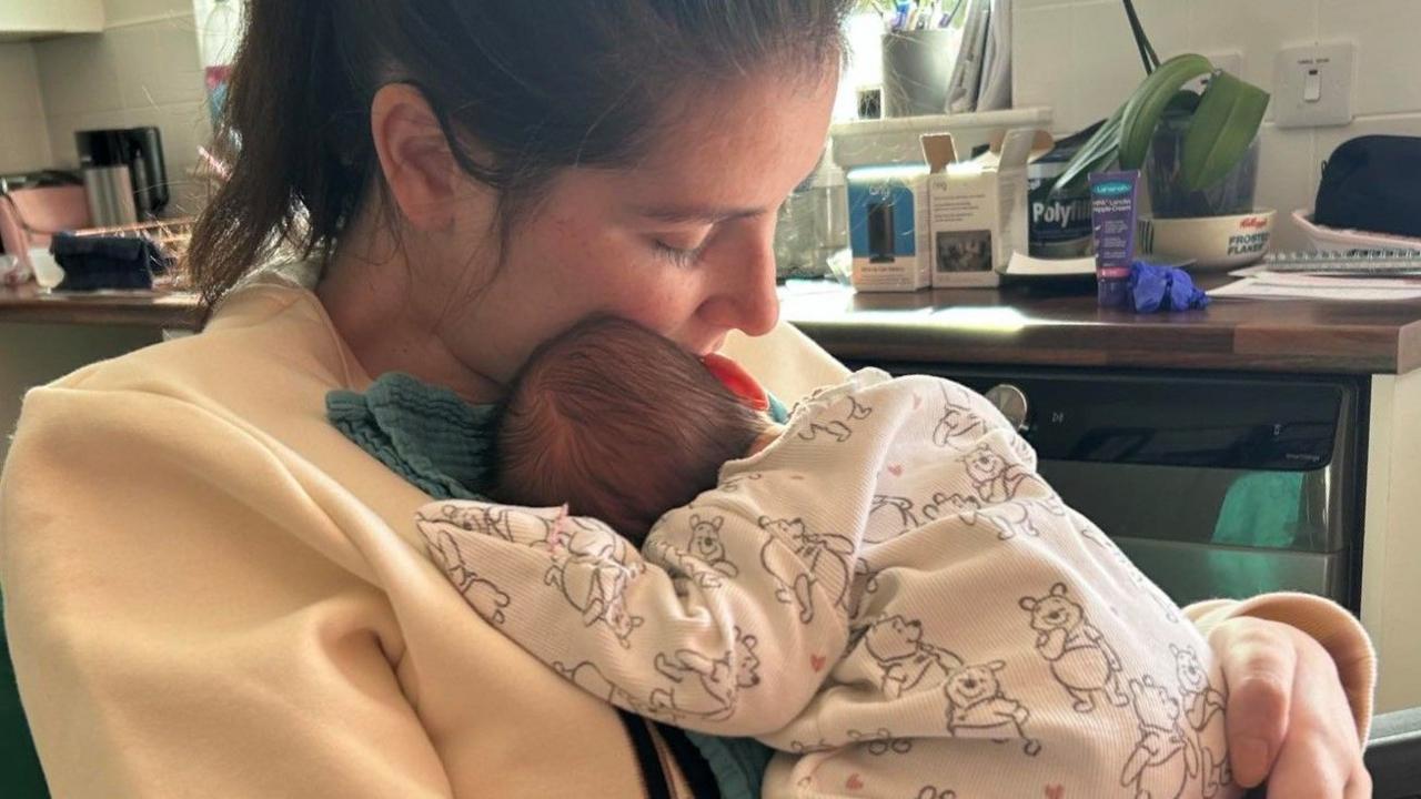 Lucy Lintott Smith holds to her chest sleeping baby SS, who is wearing a Pooh onesie, in their kitchen.