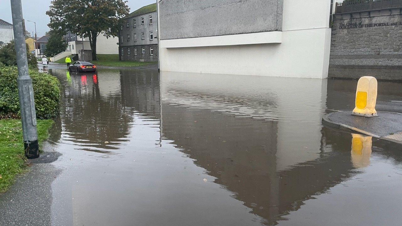 A flooded road. water us above the pavement. A car in the distance has been stopped by a person in a high visibility jacket. 