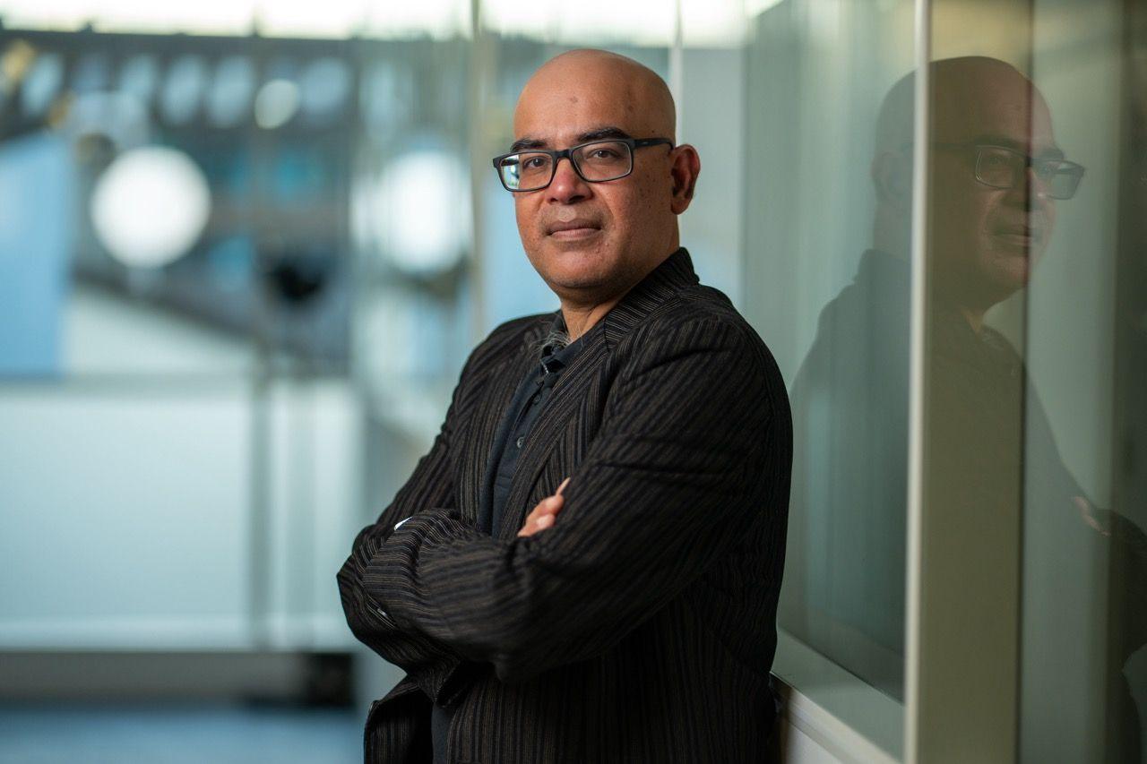 Professor Mahesh Anand stands with his arms folded and his back to a reflective screen in a corridor.