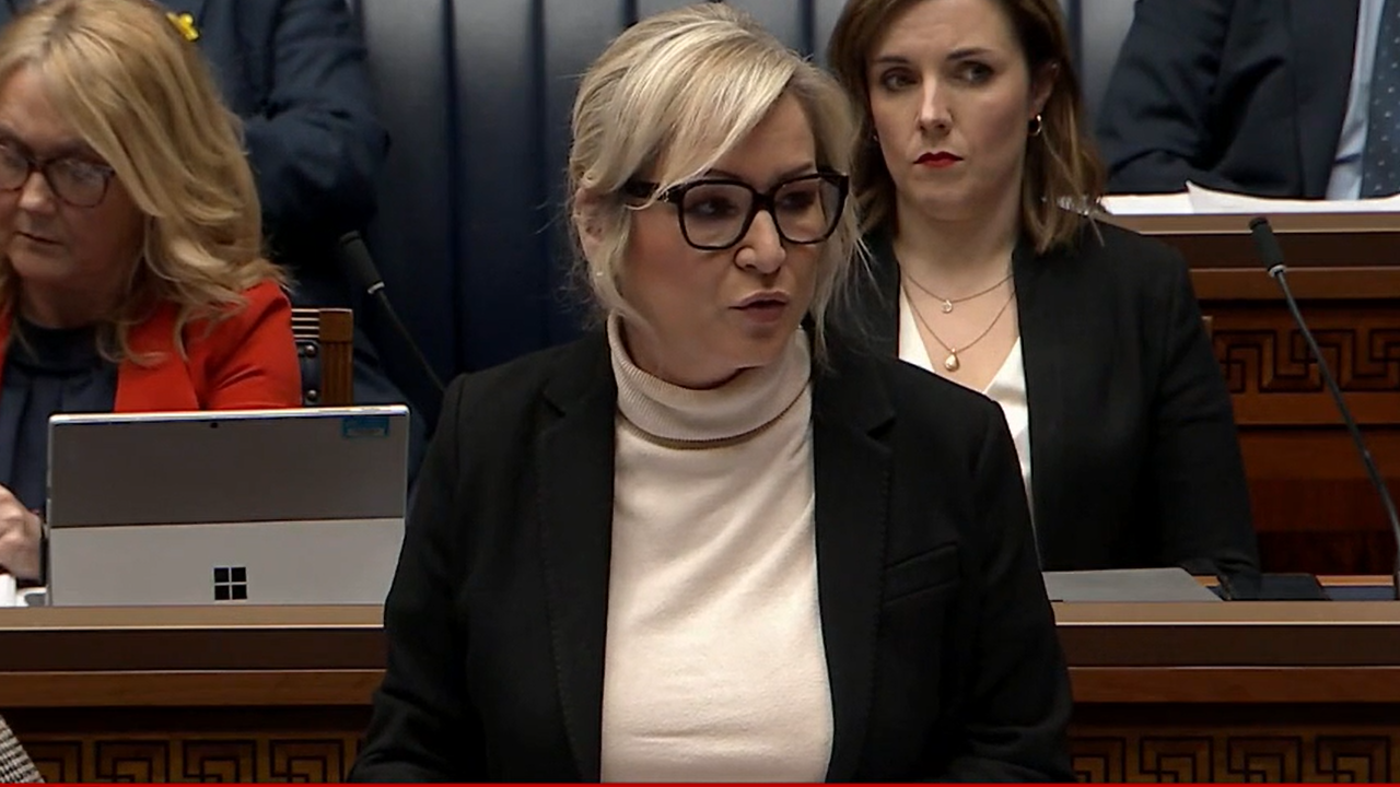 Michelle O'Neill speaking in the Stormont assembly. She has shoulder-length blonde hair and is wearing a dark jacket with light coloured top.