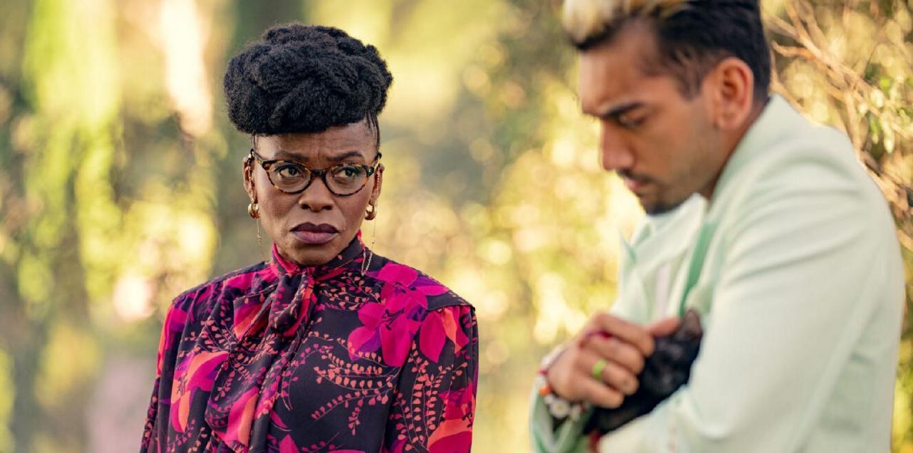 Rakie Ayola in Kaos, wearing a colourful top, glasses and a stern expression, staring at another character, a man dressed in white