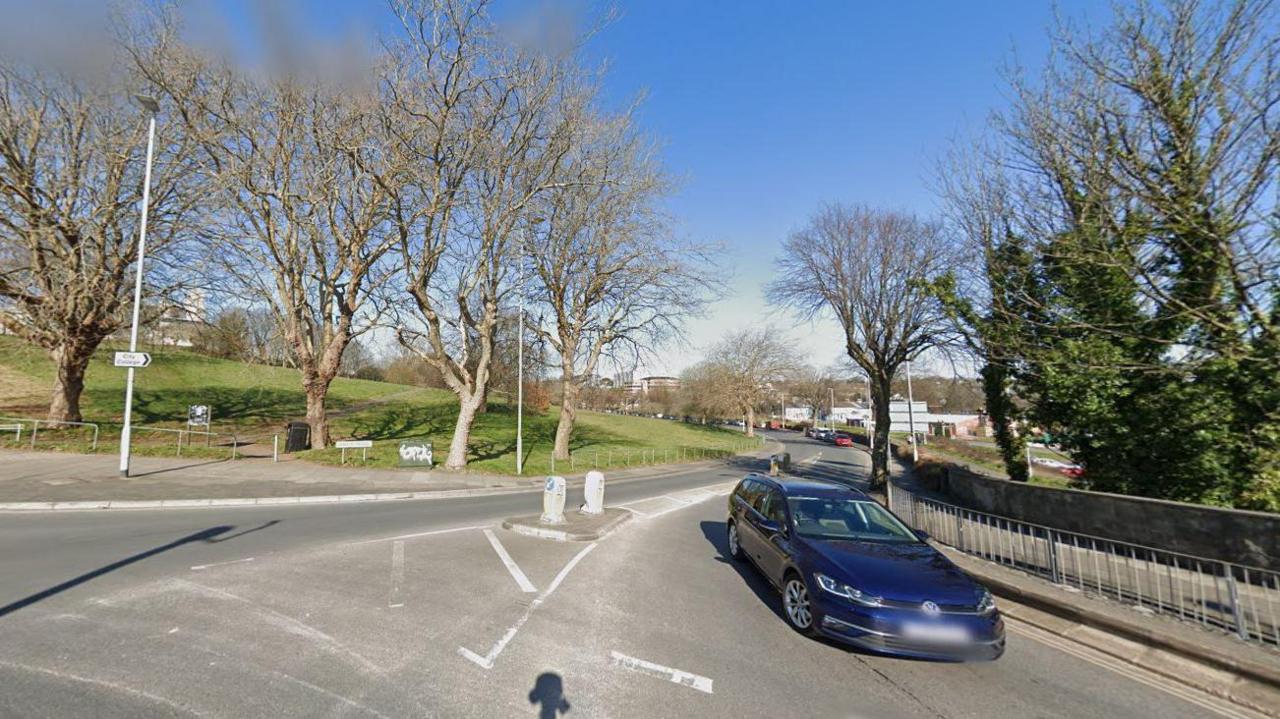 A blue car leaving Kings Road junction with has a large field to the left of it and trees either side.