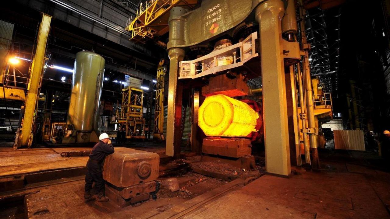 A view of the factory floor at Sheffield Forgemaster. A ma wearing overalls and a white hard hat leans on heavy machinery, while a huge machine is at work in front of him. 